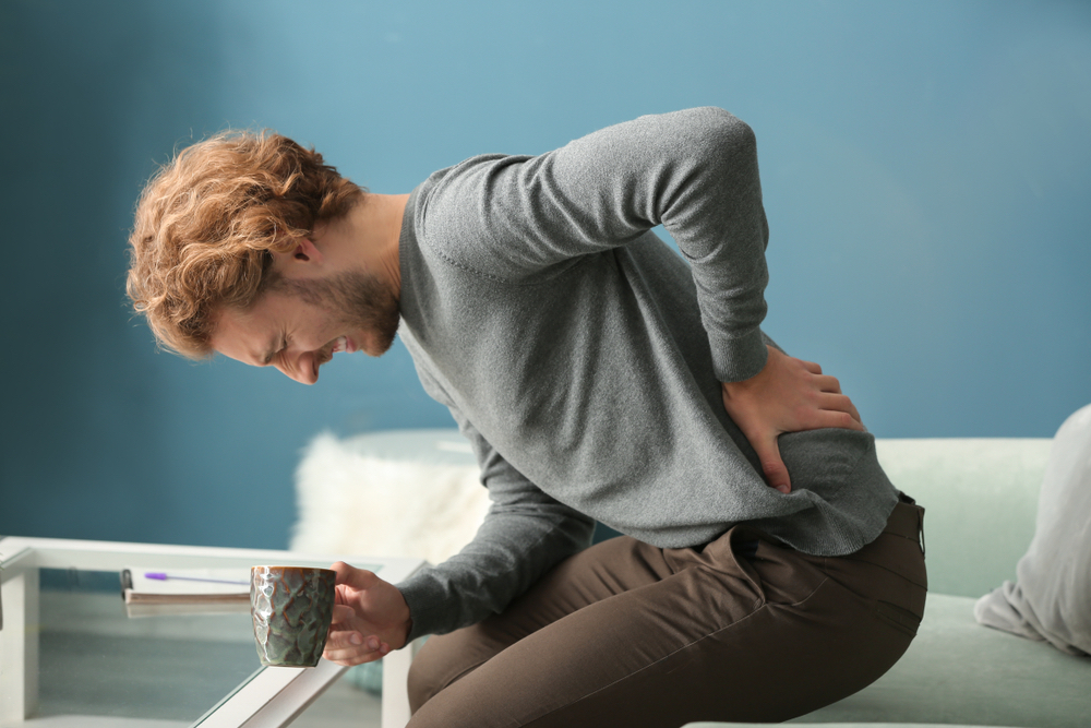 A man with sharp back pain leans forward as he sits on a couch